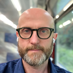 A person wearing glasses and earbuds, with bright ceiling lights and a window showing greenery outside.