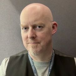 A person is shown from the shoulders up against a plain grey background, wearing a light-colored shirt, a dark vest, and a lanyard with text reading “THE UNIVERSITY OF EDINBURGH.”