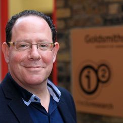 A person wearing a dark blazer over a collared shirt stands outdoors near a brick wall, with a sign in the background displaying the Goldsmiths University of London logo.