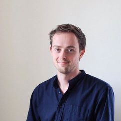 A person shown from the torso up, wearing a dark buttoned shirt, standing against a plain light-colored wall.