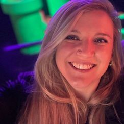 A person with long light-colored hair stands in a neon-lit environment with bright green and purple objects in the background.