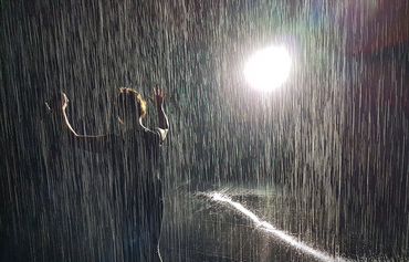 Photo of person holding hands up as if dancing in the rain with what looks like rain and a bright spotlight behind them